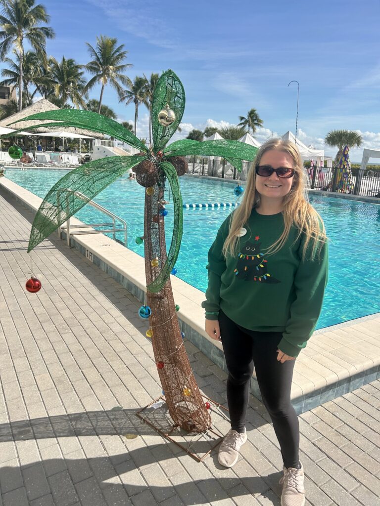 sundial staff member posing in cat christmas sweater in front of christmas palm tree and the main resort pool